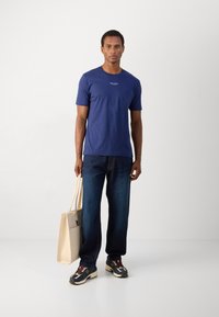 Marineblå bomull T-skjorte med rund hals. Løse jeans i mørk blå denim. Beige tote bag holdt i den ene hånden. Flerfargede joggesko.