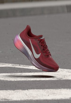 Chaussure de course Nike rouge avec le logo swoosh blanc, en l'air au-dessus de l'asphalte avec des marquages routiers blancs, de petits débris flottant à proximité.
