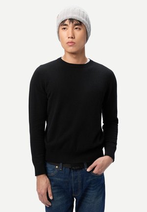 Junger Mann mit grauer Strickmütze, schwarzem Pullover und blauen Jeans, steht mit einer Hand in der Tasche vor einem einfarbigen weißen Hintergrund.