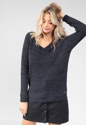 Blonde Frau mit lockerem Haar, die einen dunkelgrauen gestrickten Pullover mit V-Ausschnitt und einen schwarzen Rock mit Knopfleiste trägt, posiert mit einer Hand am Kopf.