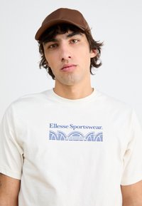 Balta medvilninė marškinėliai su mėlynu "Ellesse Sportswear" logotipu ir bangų raštu. Ruda kepurė su struktūrizuota eiga ir glotnia tekstūra.