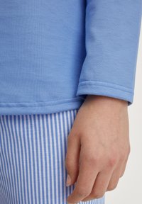 Himmelblaues Langarmoberteil mit glatter Textur, kombiniert mit blau-weiß gestreiften Hosen. Sichtbare Nähte entlang des Ärmelrandes.