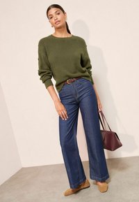 Pulover tricotat de culoare verde khaki, blugi negri cu croi larg și balerini bej. Geantă burgundă cu un design unghiular. Texturi netede și croi relaxat.