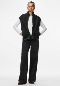 Tumši pelēka flīsa veste ar augstu apkakli, priekšā rāvējslēdzēju un lielām kabatām, uzvilkta virs baltas garroku krekla un kopā ar melnām biksēm.