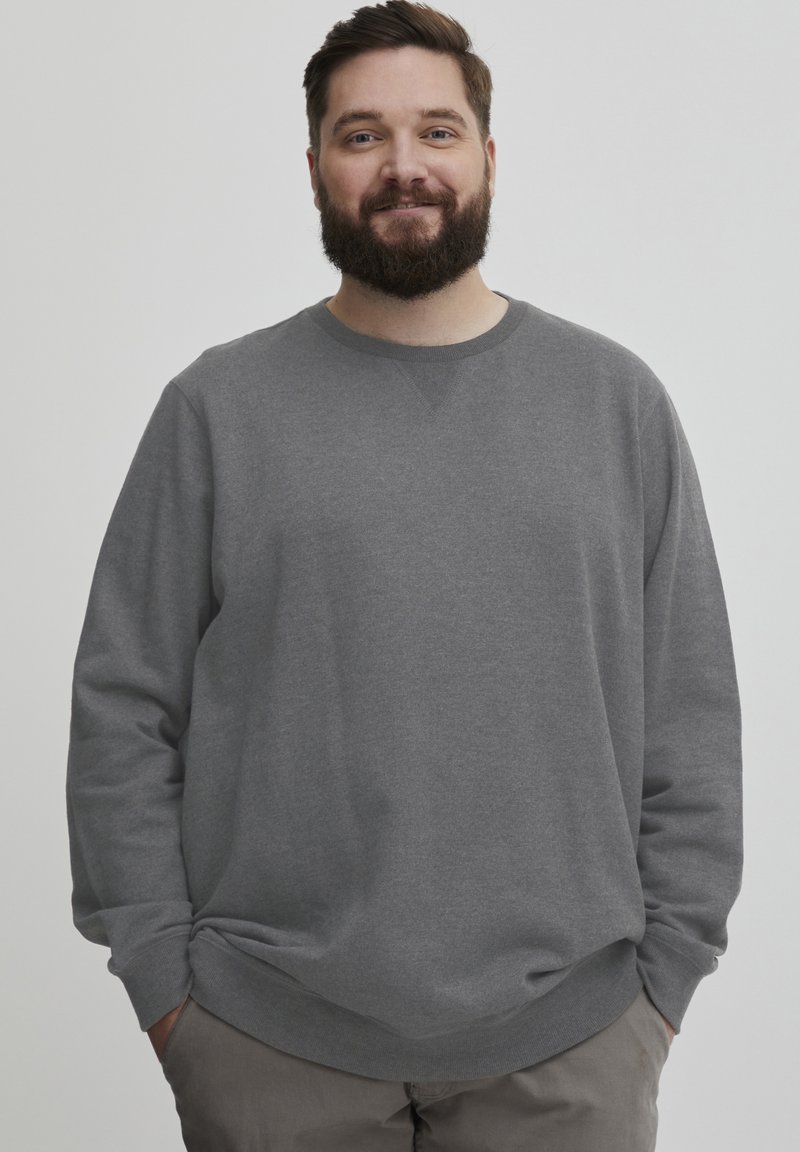 Sudadera gris con mangas largas, escote redondo, textura suave y puños y dobladillo acanalados. Llevada de forma casual, con las manos en los bolsillos, fondo neutro.