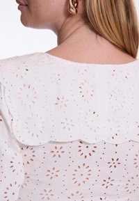 Witte blouse met ingewikkeld bloemiah-motief, gegolfde halslijn en gestructureerde stof. Beschikt over ronde oorbellen en haar tot op schouderlengte.