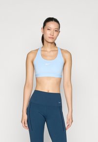 Sujetador deportivo atlético azul claro con tirantes finos, que presenta un pequeño logo blanco en la parte frontal, combinado con leggings oscuros de cintura alta.