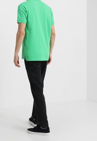 Grön bomullspiké med korta ärmar, kombinerad med svarta byxor och mörka sneakers; bakifrån, slätt tyg, standardpassform.