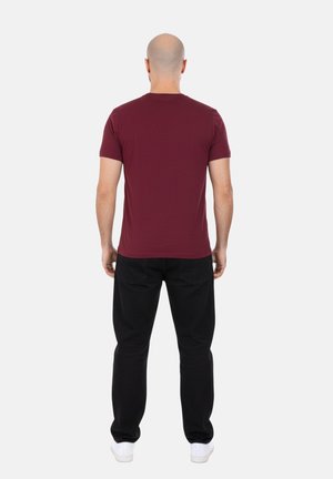 Uomo con testa rasata che indossa una t-shirt bordeaux a maniche corte tinta unita, pantaloni neri e scarpe da ginnastica bianche, visto di spalle.