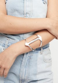 Brazalete de metal plateado con un diseño moderno y elegante, que presenta una forma abierta y curva y una textura suave, llevado en un brazo descubierto.