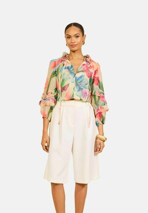 Nő színes virágos blúzt visel fodros ujjakkal és fehér, magas derekú térdig érő rövidnadrágot, arany karkötőkkel és fülbevalókkal kiegészítve.