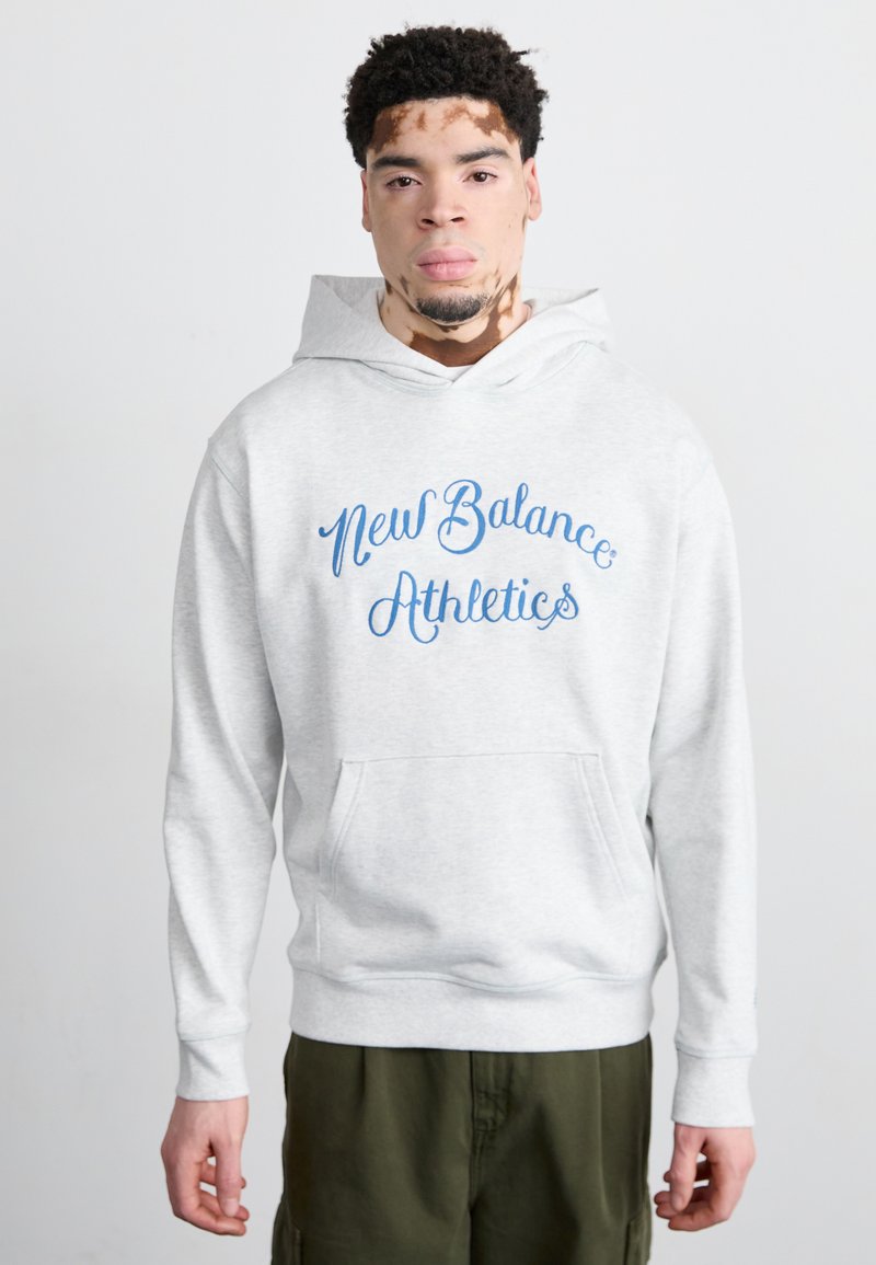 Siva kapuca s prednjo kenguru žepom, koja sadrži plavi tekst "New Balance Athletics". Tkanina izgleda mekano s opuštenim krojem i podesivom kapuljačom.