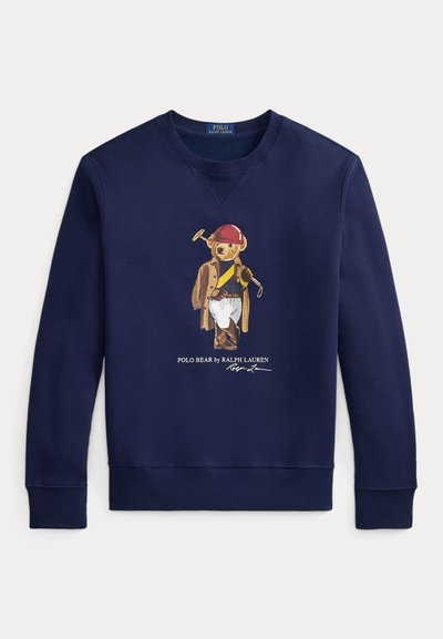 Marineblå genser med en bjørn som har på seg en rød hjelm, brun kåpe, hvite bukser og holder en polo-kølle, med teksten "Polo Bear by Ralph Lauren" under.