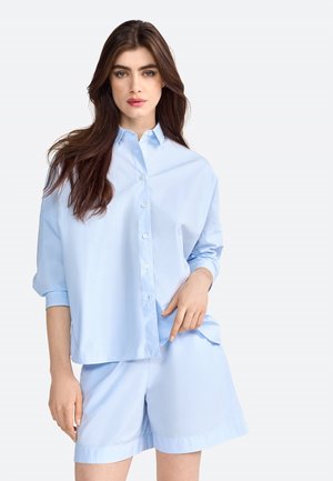 Camicia azzurro chiaro con maniche arrotolate, abbinata a pantaloni corti azzurro chiaro. Tessuto morbido e vestibilità rilassata, con una superficie liscia.