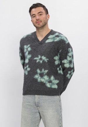 Man draagt een donkergrijze pluizige trui met lichtgroene bloemmotieven en lichtgrijze spijkerbroek, staand met handen achter de rug.