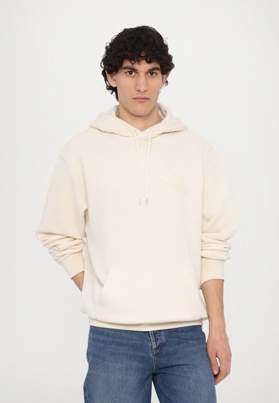 Hoodie aus cremefarbenem Baumwollstoff mit einer vorderen Tasche und gerippten Bündchen; mit aufgesticktem Text in einer kontrastierenden Farbe auf der Brust.