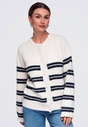 Femme portant un cardigan blanc boutonné avec des rayures horizontales noires et un jean bleu, debout devant un fond clair uni.
