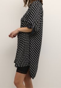 Femme portant une longue chemise noire et blanche à motifs avec les manches retroussées et un short noir orné de dentelle, sur un fond uni.