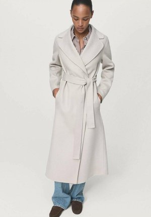 Manteau en laine gris clair avec un design enveloppant, manches longues, grands revers, et une ceinture assortie. Porté par-dessus une chemise rayée et un jean bleu.