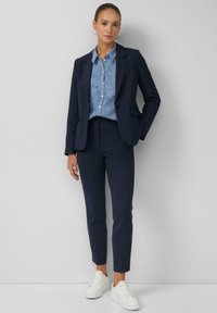 Marineblauer Blazer und passende Hose, kombiniert mit einem hellblauen, strukturierten Hemd. Weiße Sneaker, minimalistische Gestaltung, maßgeschneiderte Passform, klassischer Look.
