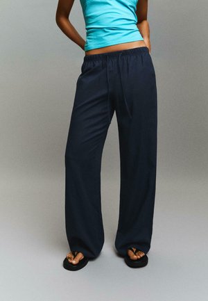 Osoba nosząca luźne granatowe spodnie z elastycznym pasem i sznurkiem, turkusowy top bez rękawów oraz czarne klapki, stojąca na szarym tle.