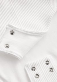 Gros plan d’un tissu blanc côtelé avec des boutons-pression argentés sur les poignets réglables et les bords du vêtement.