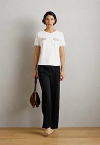 T-shirt bianco a maniche corte con tasche decorative, abbinato a pantaloni larghi neri. Il modello tiene una borsa marrone, in piedi su un pavimento in legno.