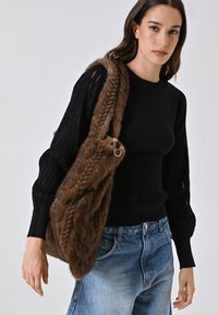 Braune Faux-Fur-Handtasche mit geflochtener Textur, versehen mit goldfarbenen Hardware-Elementen. Das Model trägt ein schwarzes Langarmoberteil und Jeans.