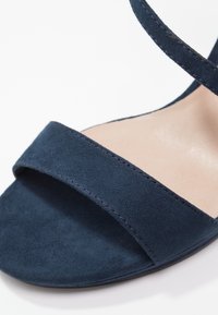 Sandale à talon haut en daim bleu marine avec une large bande avant et une fine bride à la cheville, présentant une texture lisse et un design minimaliste.