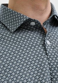 Cămașă cu model geometric în nuanțe de bleumarin și alb, având un guler cu nasturi și o textură contrastantă a materialului. Două nasturi sunt vizibili.