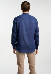 Granatowa lniana koszula z kołnierzykiem stójkowym i długimi rękawami, z zaokrąglonym dołem i zapięciem na guziki przy mankietach. Materiał ma subtelną teksturę.