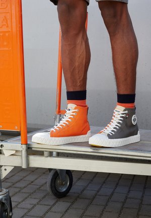 Person wearing mismatched high-top sneakers, one orange and one gray, standing on a metal platform with wheels on a paved surface.