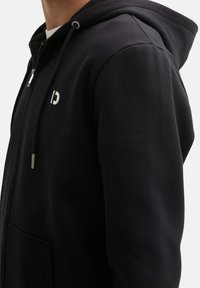 Felpa nera con zip, dotata di tasca a marsupio, cappuccio regolabile e un piccolo logo bianco sul petto. Tessuto morbido e vestibilità casual.