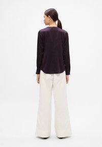Dunkelviolettes Langarm-Shirt mit geradem Schnitt, glatter Textur und geknöpften Manschetten, kombiniert mit hoch taillierten, cremefarbigen Weitbein-Hosen.