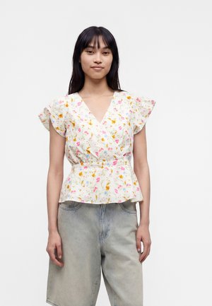 Femme aux longs cheveux foncés portant un chemisier blanc à manches courtes fleuries et un short en denim gris clair, debout devant un fond uni.