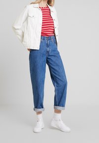 Person med en vit jeansjacka över en röd och rosa randig skjorta, blå uppvikta jeans, vita strumpor och vita sneakers som står mot en grå vägg.