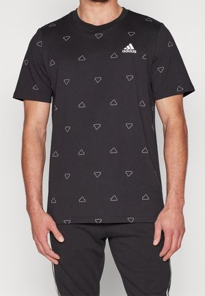Homme portant un t-shirt noir Adidas avec de petits motifs triangulaires blancs, accompagné d'un pantalon noir avec des bandes blanches sur les côtés.