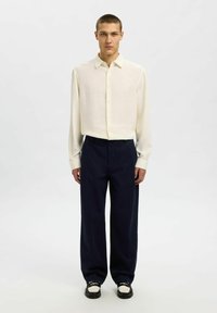 Camicia button-up beige chiaro con maniche lunghe, abbinata a pantaloni a gamba larga blu scuro e scarpe nere con riflessi bianchi.