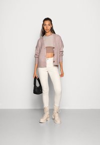 Cardigã de malha rosa claro com botões de madeira sobre um top cropped bege. Jeans skinny brancos e botas bege com atacadores. Bolsa trançada preta.