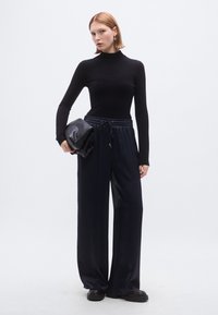 Melns garroku tops ar augstu apkakli, kombinēts ar tumši ziliem plaša piegriezuma biksēm. Modelis tur tumšu rokassomiņu un valkā masīvas melnas kurpes.