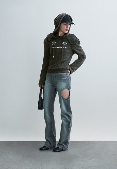 Άτομο που φοράει σκούρα γκρι φούτερ Off-White, μπλε τζιν με ανοίγματα στον μηρό, μαύρο καπέλο και κρατώντας μια μικρή μαύρη τσάντα, στέκεται σε γκρίζο δάπεδο.