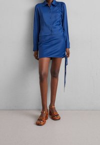 Blaue Satinbluse mit Knopfverschluss und seitlichem Raffung, kombiniert mit einem passenden blauen Minirock. Das Modell trägt orangefarbene Schuhe mit Schlangenprint und Absatz.