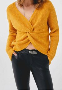 Femme portant un pull en maille torsadée jaune moutarde et un pantalon en cuir noir avec ceinture à boucle argentée, mains dans les poches, sur un fond uni.