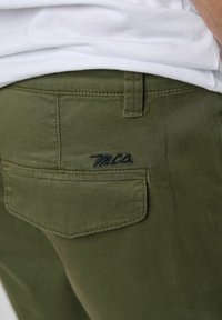Pantalones verde oliva con una textura suave. Cuenta con un bolsillo trasero con un logo bordado y detalles de costura visibles.