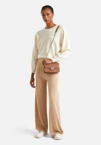 Maglione beige a maglia, pantaloni crema e una borsa a tracolla marrone con hardware argentato. La modella è in piedi davanti a uno sfondo neutro.