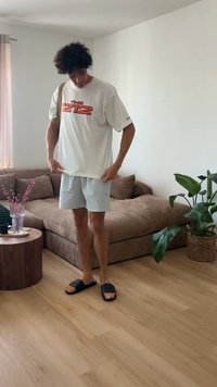 Νέος άνδρας με λευκό γραφικό μπλουζάκι, ανοιχτόχρωμο σορτς και μαύρα σανδάλια στέκεται σε ένα σαλόνι με καφέ καναπέ, ξύλινο τραπέζι και γλαστράκι.
