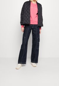 Giacca bomber trapuntata nera su una felpa rosa, abbinata a pantaloni cargo in denim scuro a gamba larga. Sneakers bianche completano l'outfit.
