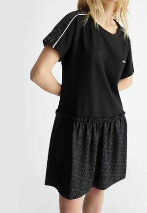 Mujer vestida con un vestido negro de manga corta con falda estampada fruncida y paneles estampados en los hombros con detalles de ribete blanco.