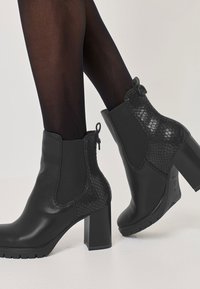 Černé kožené kotníkové boty s texturovaným hadím vzorem po stranách, na blokovém podpatku, nošeny s průhlednými černými punčochami na bílé podlaze.