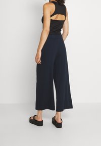 Zwarte mouwloze top met een open rug, gecombineerd met een brede, geribbelde marineblauwe broek en zwarte platform sandalen. Eenvoudig, ontspannen ontwerp.